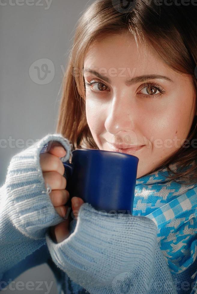 una mujer joven y triste que se queda en casa debido a la gripe y la temperatura en un suéter cálido y una bufanda con una taza de té cerca de la ventana foto