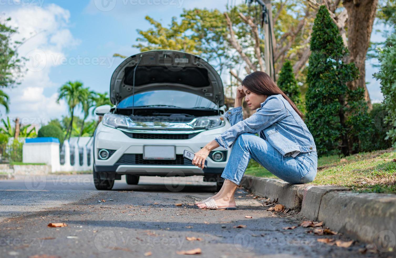 Asian woman sitting beside car after a car breakdown on street. Concept of vehicle engine problem or accident and emergency help from Professional mechanic photo