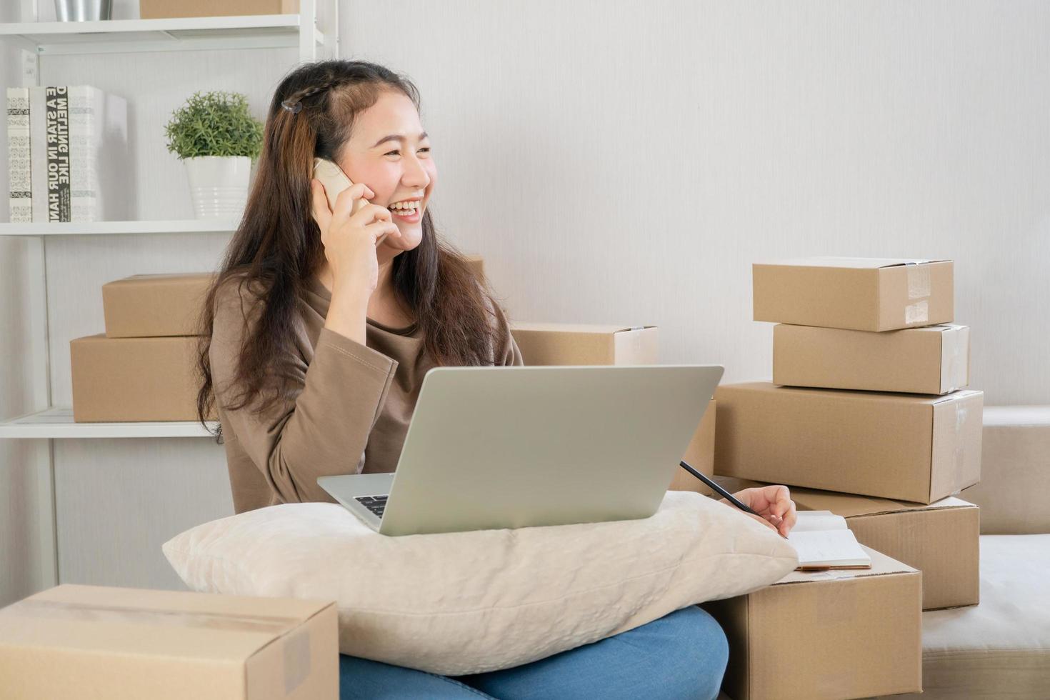 Happy Asian young entrepreneur, talking on the smartphone with customer for tack order, She smile for sales success at home office, Concept of merchant business online and eCommerce photo