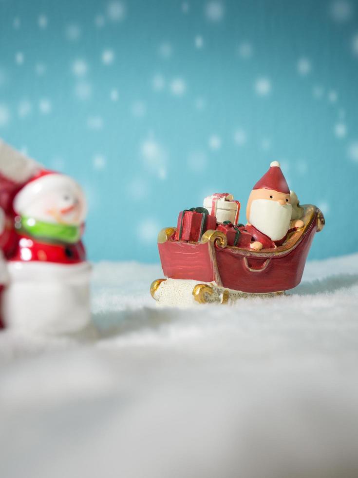 feliz santa claus con caja de regalos en el trineo de nieve yendo a casa. cerca de la casa hay un muñeco de nieve y un árbol de navidad. santa claus y casa en la nieve. concepto de navidad y feliz año nuevo. foto