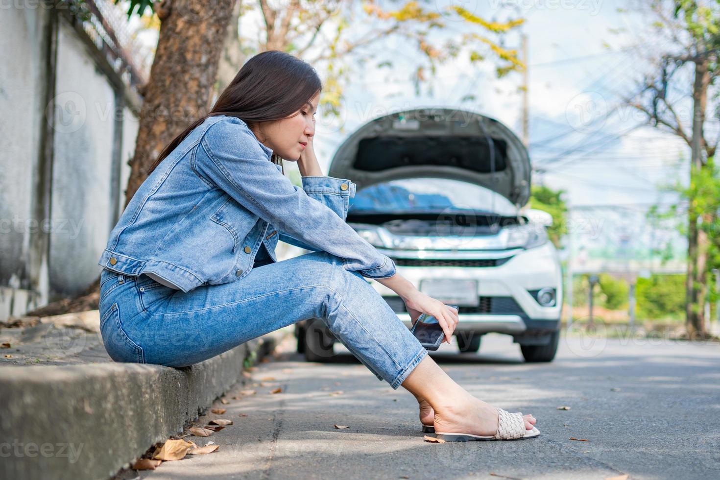 Asian woman sitting beside car after a car breakdown on street. Concept of vehicle engine problem or accident and emergency help from Professional mechanic photo