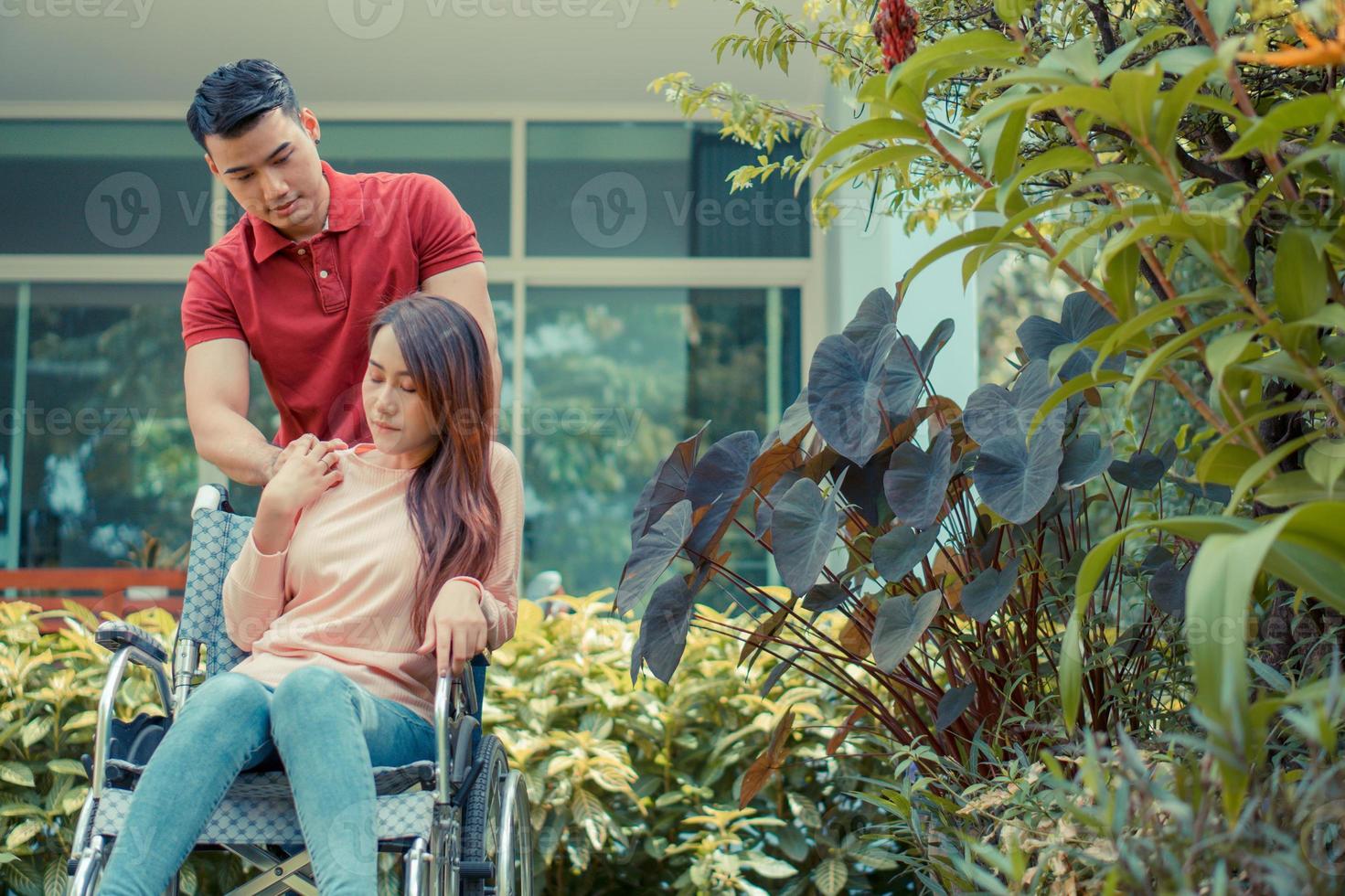 mujer asiática en silla de ruedas e infeliz y dolorosa. un hombre parado detrás de la silla de ruedas y está animando a su esposa, cuyos pies le duelen la pierna debido a un accidente. concepto de cuidado y apoyo foto