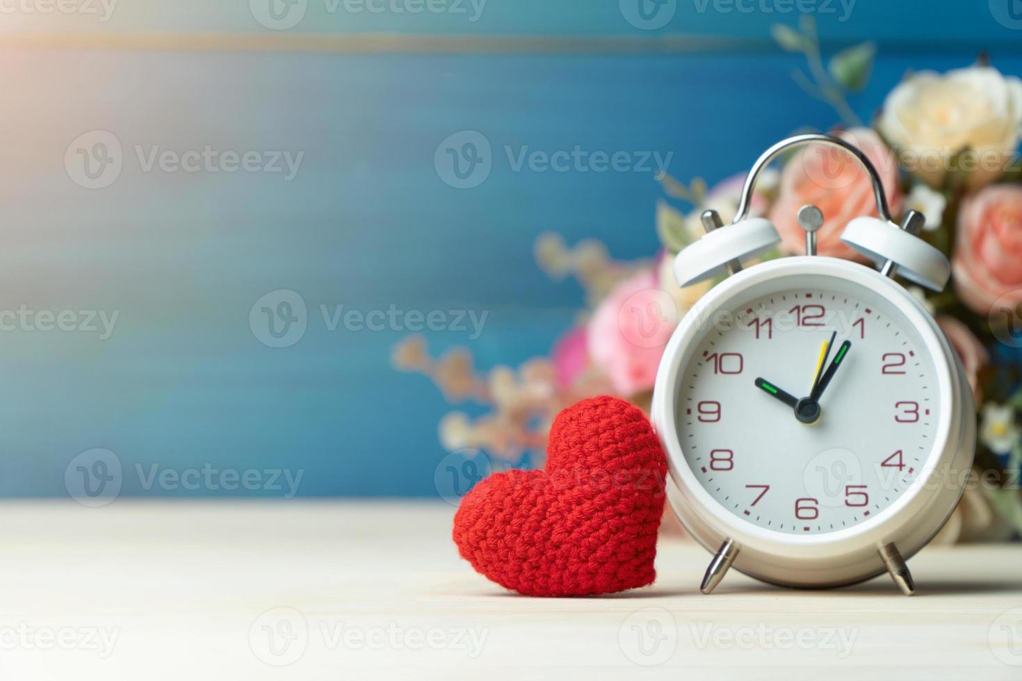 Valentines day concept. hand make yarn red heart beside white alarm clock in front of rose flower bouquet on wooden table and blue background photo