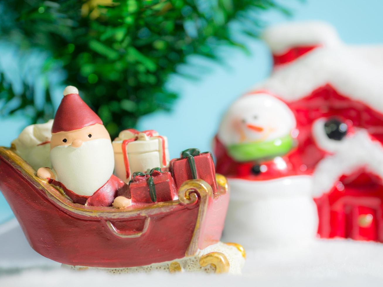 feliz santa claus con caja de regalos en el trineo de nieve yendo a casa. cerca de la casa hay un muñeco de nieve y un árbol de navidad. santa claus y casa en la nieve. concepto de navidad y feliz año nuevo. foto