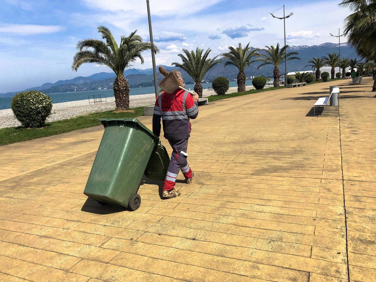 A male worker cleaner in uniform, a robe holding a spade on his shoulder and dragging a trash can on wheels on a tropical warm paradise resort against the backdrop of green palm trees photo