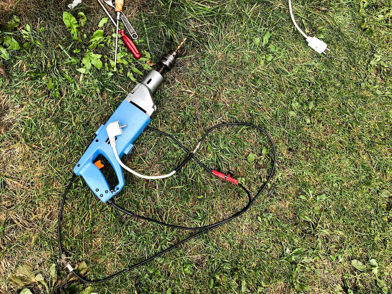 a drill with a long drill inside lies on the grass. renovation in nature. next to a hacksaw, hammer and other tools. the drill is connected through a white extension cord. building a village house photo