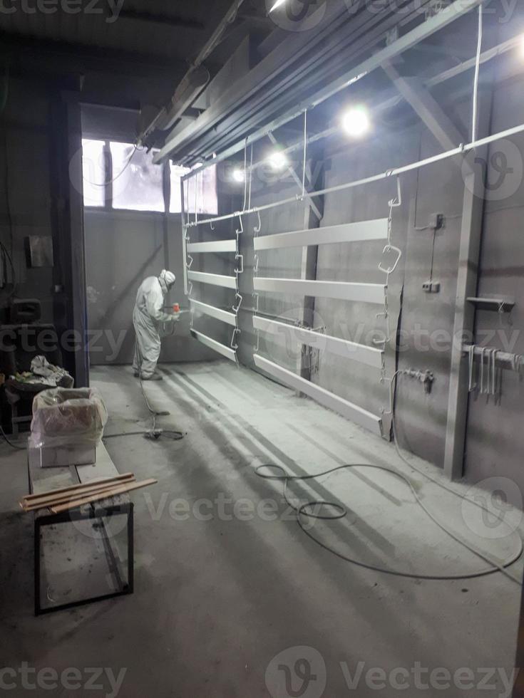 A male painter in a white disposable protective suit paints a metal structure with a spray gun at a paint factory photo