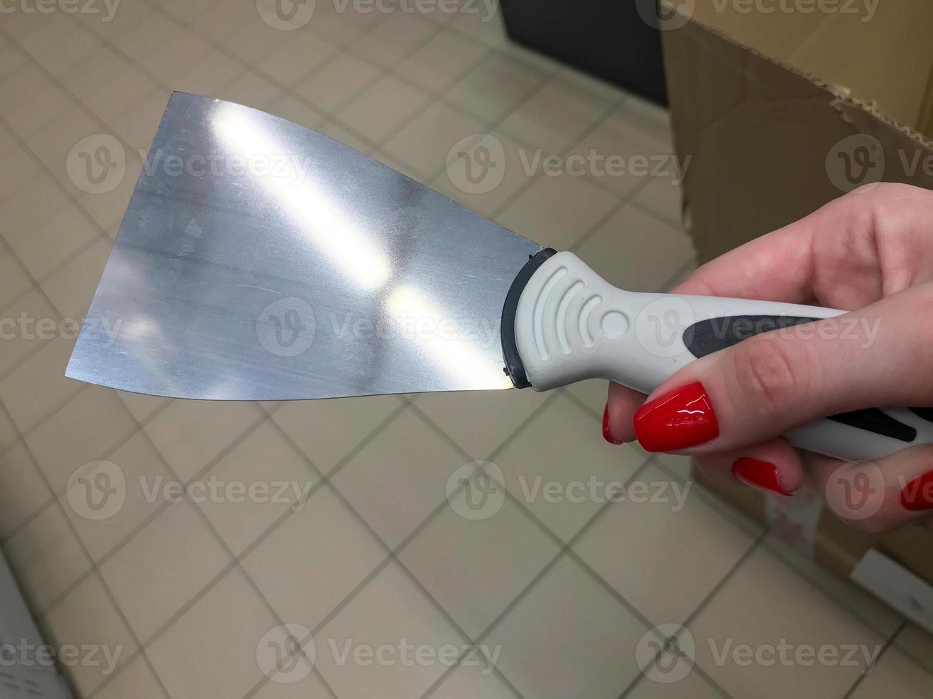 raspador para aplicar masilla, yeso. trabajos de pintura. reparación casera de bricolaje. un raspador en manos de una mujer con una manicura roja brillante. raspador con punta de metal y mango de plástico foto