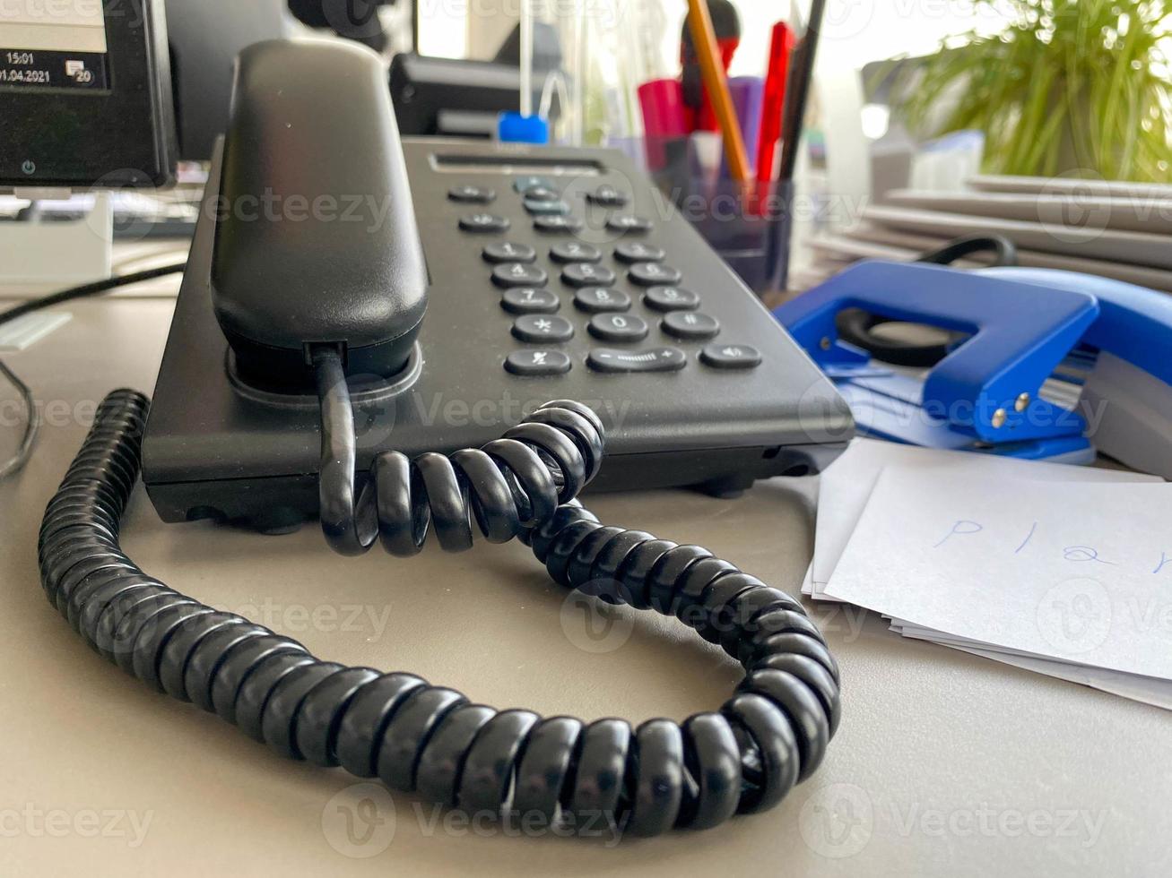 Black landline telephone with a tube, buttons and a wire on the work table at the office desk with office supplies. Business work photo