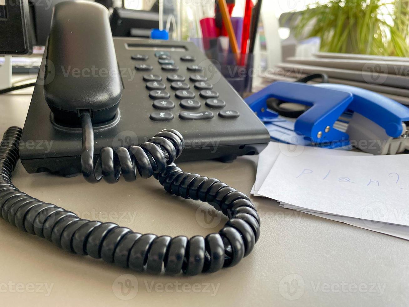 Black landline telephone with a tube, buttons and a wire on the work table at the office desk with office supplies. Business work photo