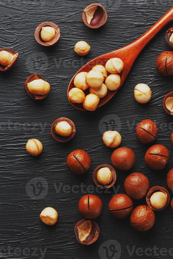 Shelled macadamia nut and peeled macadamia nut on a black textural background in a wooden spoon. Low contrast photo