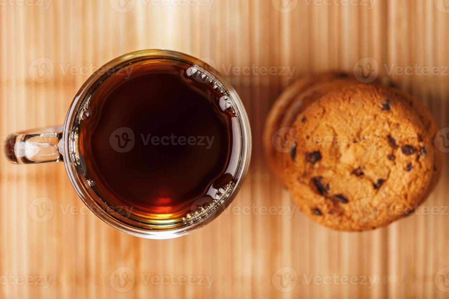 un montón de galletas de avena con trocitos de chocolate y una taza de fragante té negro caliente sobre un sustrato de bambú, sobre un fondo oscuro. Galletas artesanales para un desayuno saludable. foto