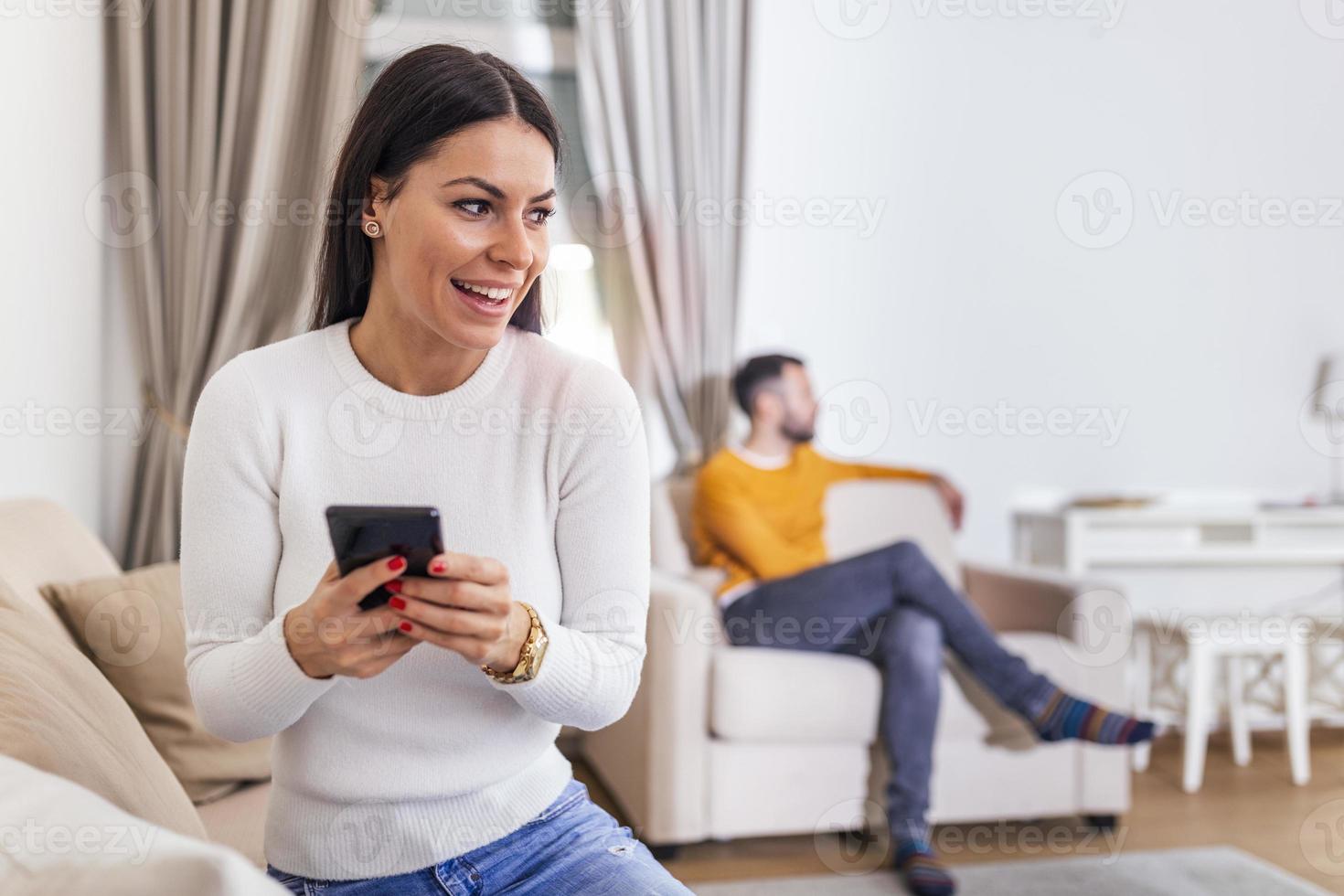 Wife turned her back to husband, reading message on phone from her lover, Man lying on sofa in the back. Cheating and infidelity concept photo