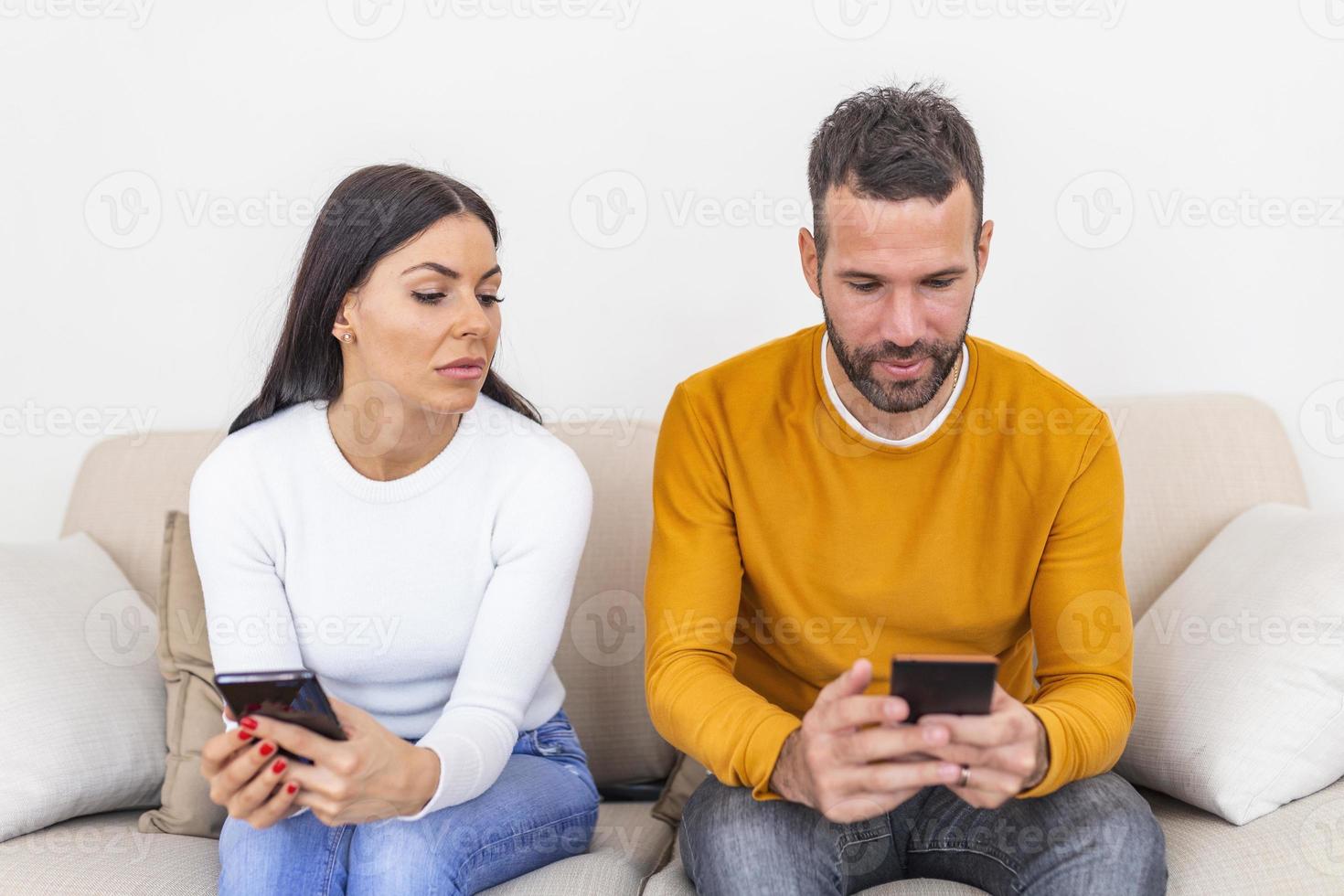 Jealous young woman with smartphone looking at smiling boyfriend using smartphone at home, relationship problem concept. Mistrust concept photo