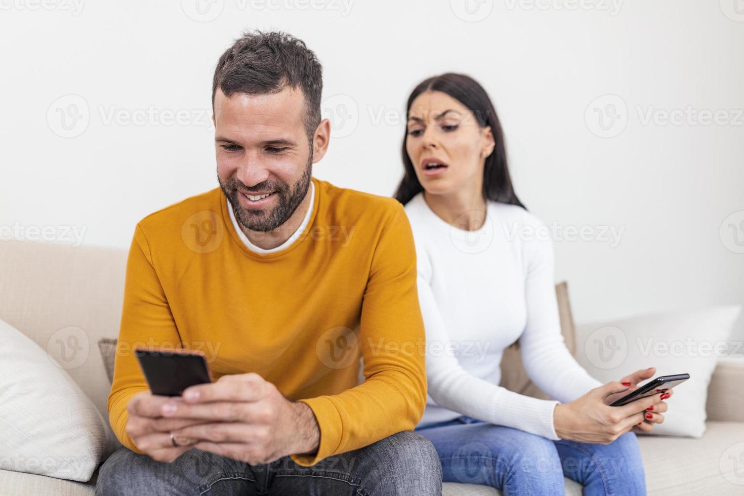 happy smiling man turned his back to wife, reading message on phone from his lover, worried woman sitting next to him, trying to peek at screen. Cheating and infidelity concept photo