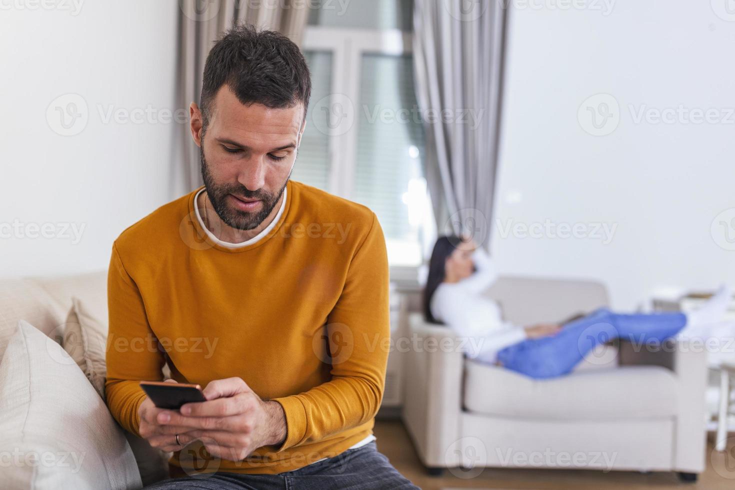 Husband turned his back to wife, reading message on phone from his lover, Woman lying on sofa in the back. Cheating and infidelity concept photo