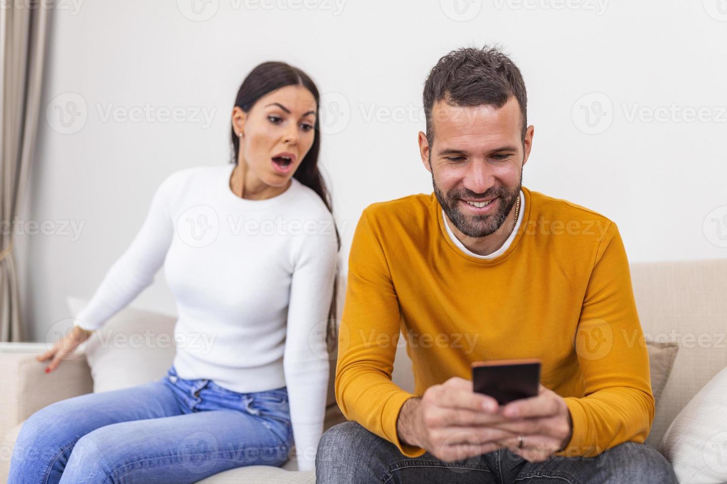 Jealous young woman with smartphone looking at smiling boyfriend using smartphone at home, relationship problem concept. Mistrust concept photo