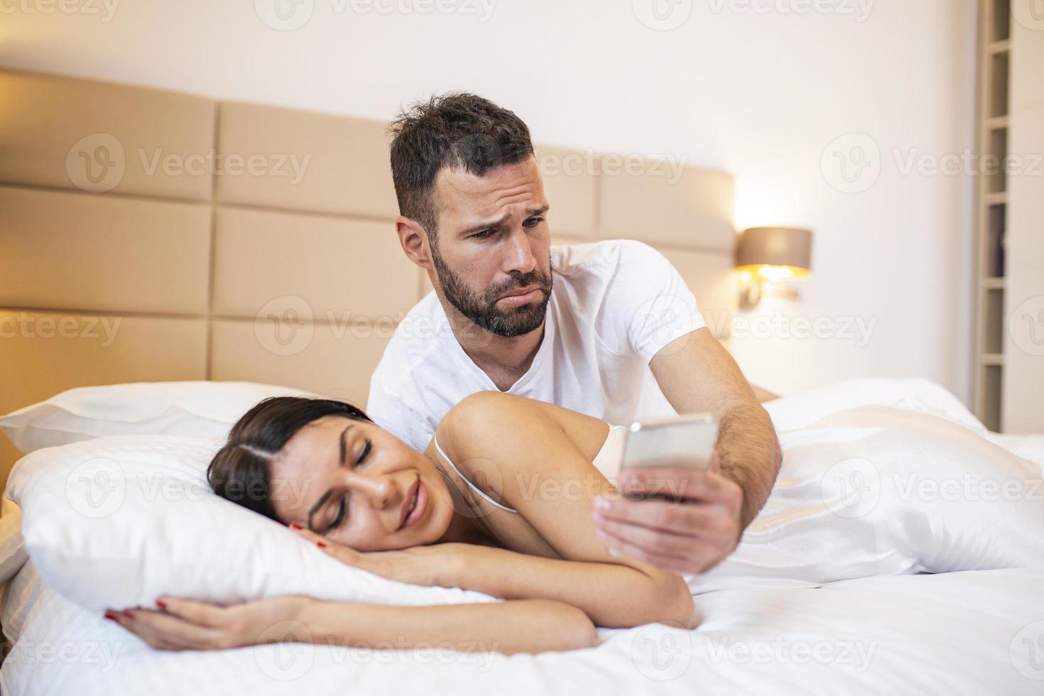 Jealous husband spying the phone of his partner while she is sleeping in a bed at home. Shocked jealous husband spying the phone of his wife while she sleeping in bed at home photo