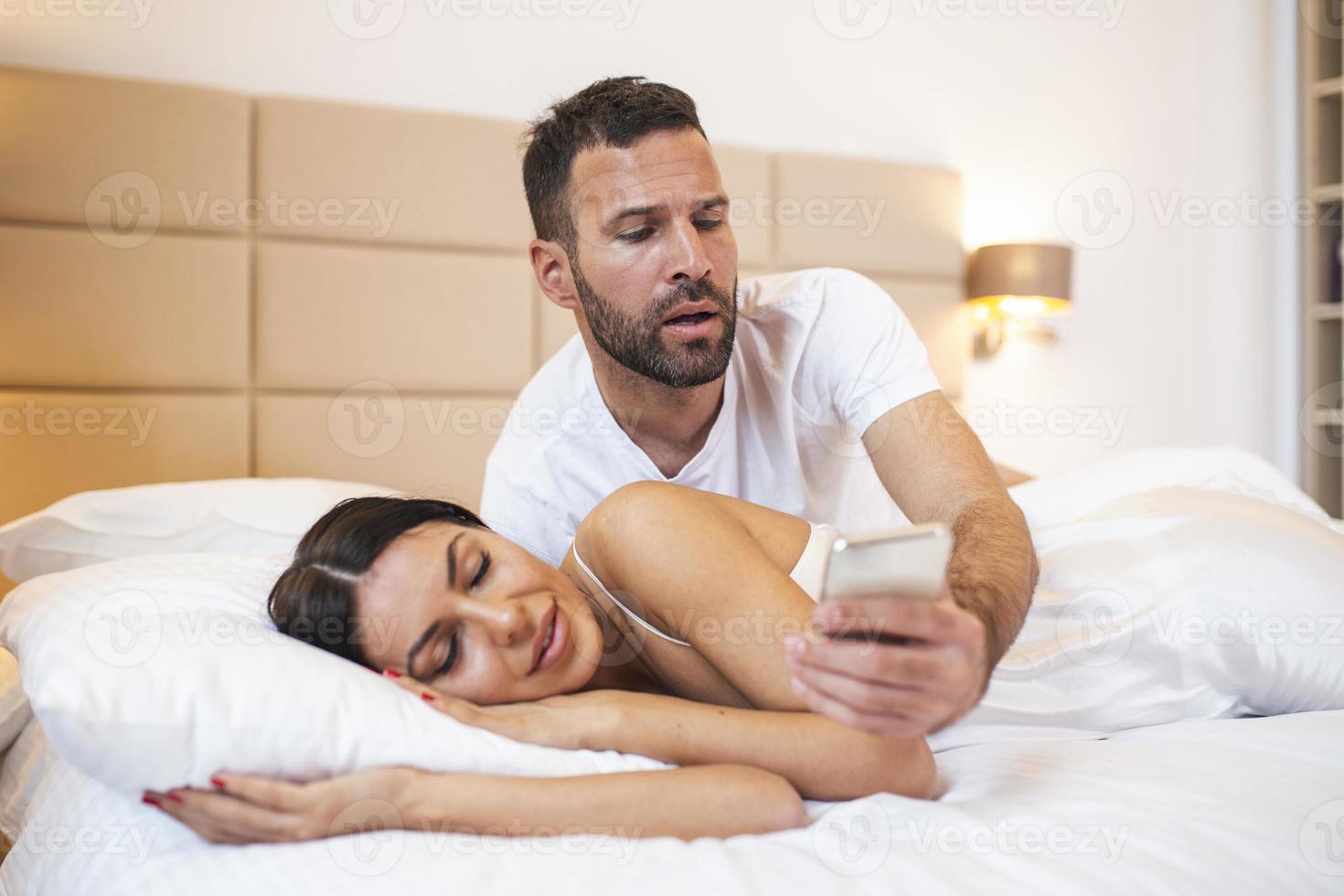 Jealous husband spying the phone of his partner while she is sleeping in a bed at home photo