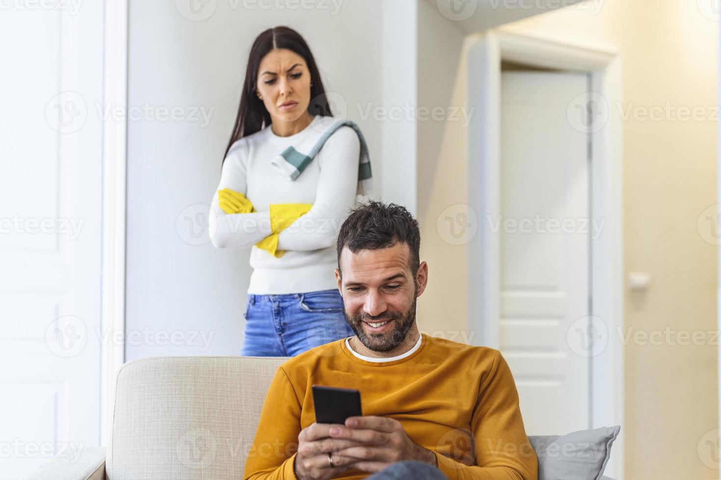 Jealous girlfriend tries to peep at boyfriends smart phone, feels sad as he texts with someone, dressed in casual sweaters, have troubles with relationships. photo