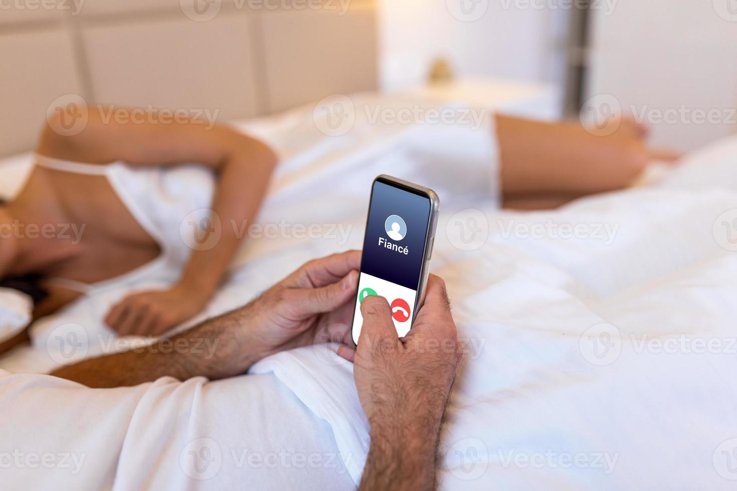 Cheating unfaithful man lying with mistress in hotel bed. Call from finace to mobile phone. Cheater having affair with secret lover and relationship with another woman. Infidelity and love triangle. photo