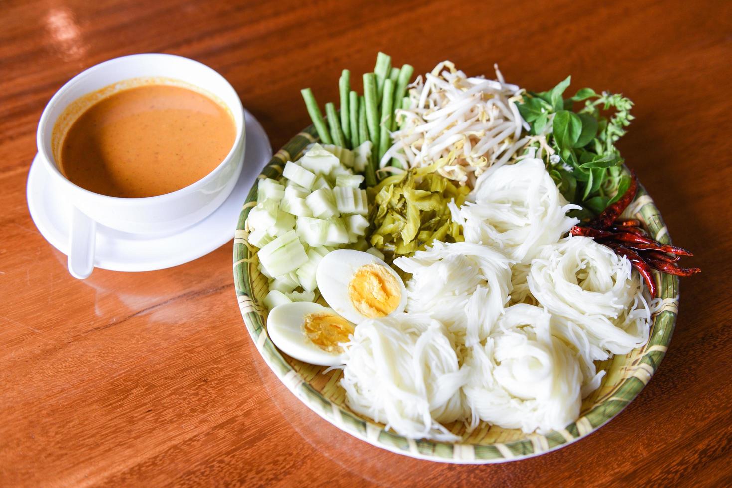 Fideos de arroz tailandeses: cierre la comida de Tailandia fideos vermicelli huevos cocidos y verduras frescas en un plato con un tazón de sopa de curry servido en una mesa de madera foto