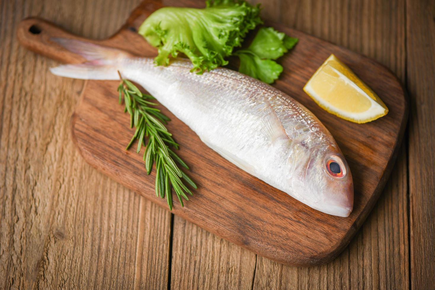 pescado crudo fresco con ingredientes limón romero y lechuga para hacer comida cocinada en tabla de cortar de madera - besugo marisco pescado congelado foto