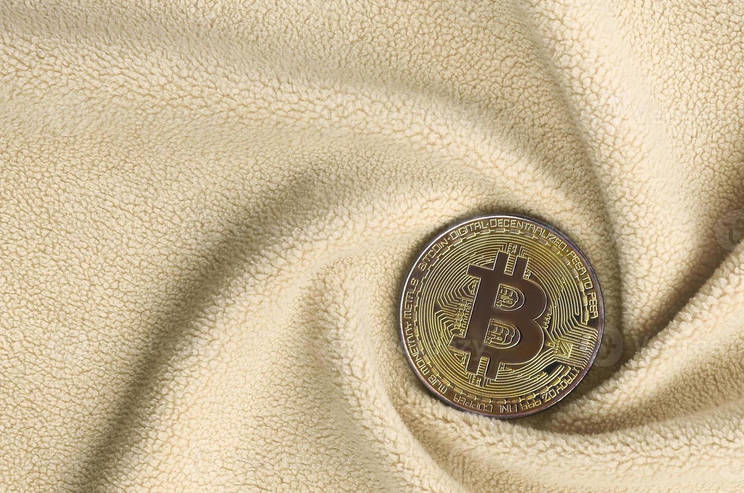 The golden bitcoin lies on a blanket made of soft and fluffy light orange fleece fabric with a large number of relief folds. The shape of the folds resembles a fan from a video card cooler photo