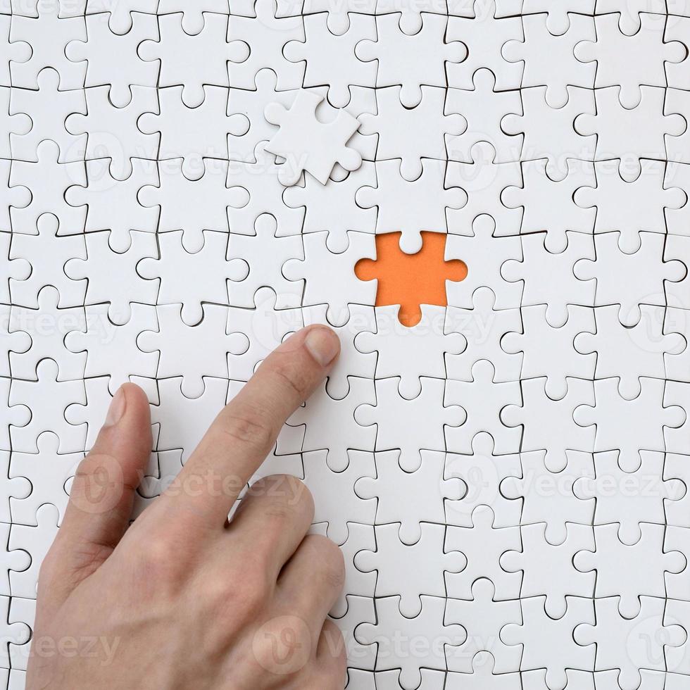The texture of a white jigsaw puzzle in the assembled state with one missing element, forming an orange space, pointed to by the finger of the male hand photo