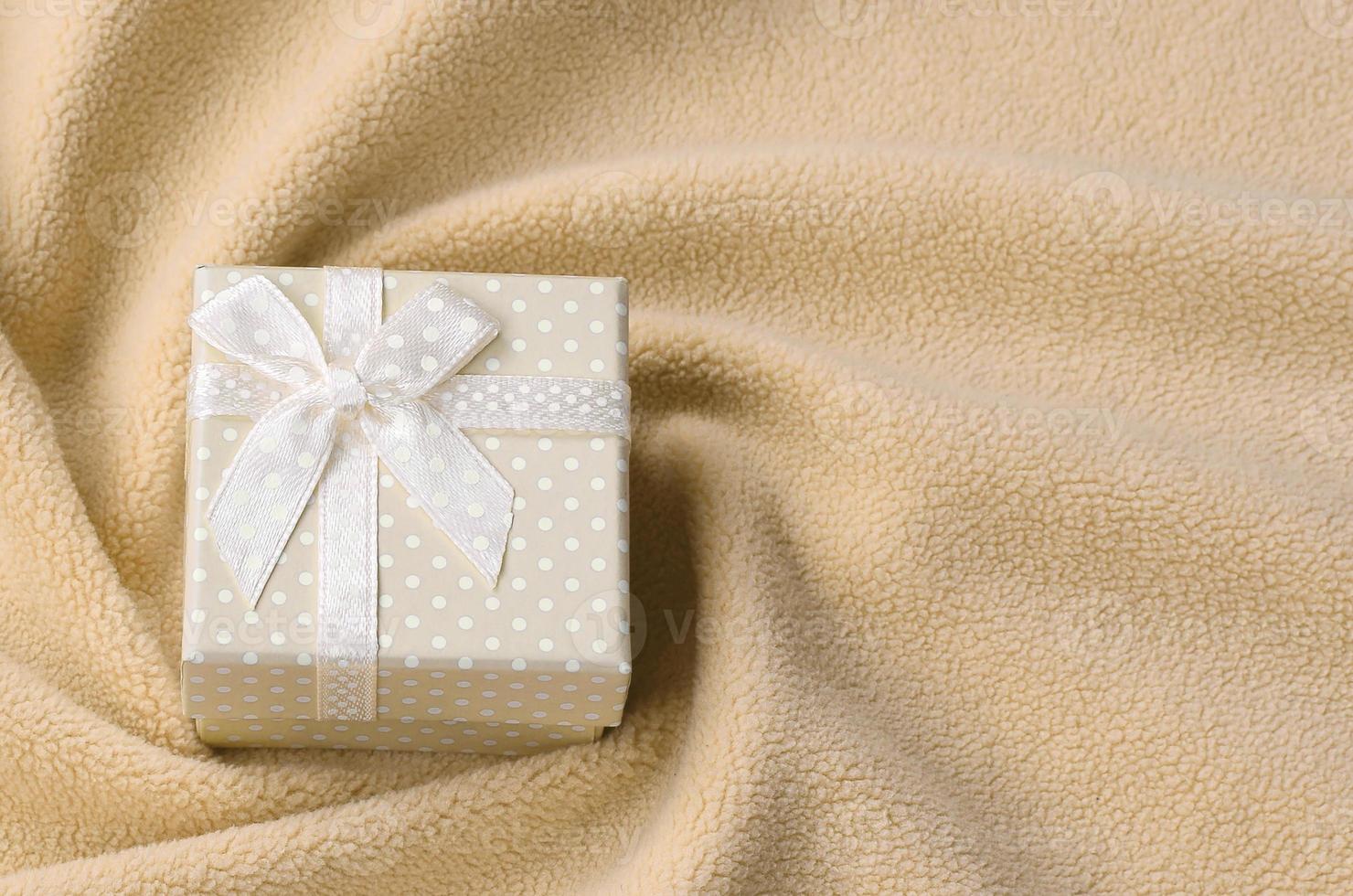A small gift box in orange with a small bow lies on a blanket of soft and furry light orange fleece fabric with a lot of relief folds. Packing for a gift to your lovely girlfriend photo