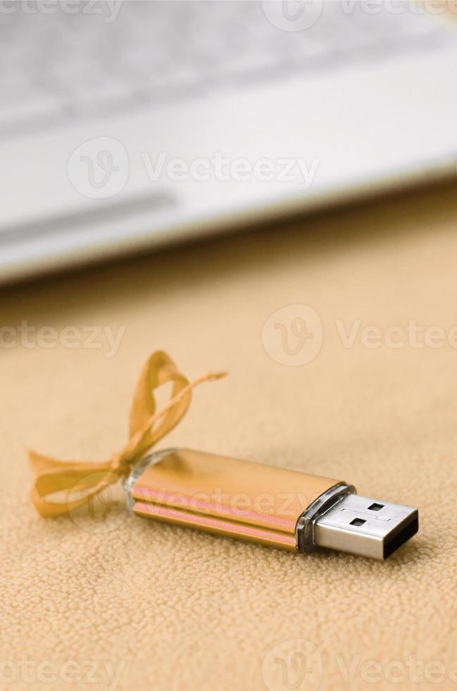 Orange usb flash memory card with a bow lies on a blanket of soft and furry light orange fleece fabric beside to a white laptop. Classic female gift design for a memory card photo