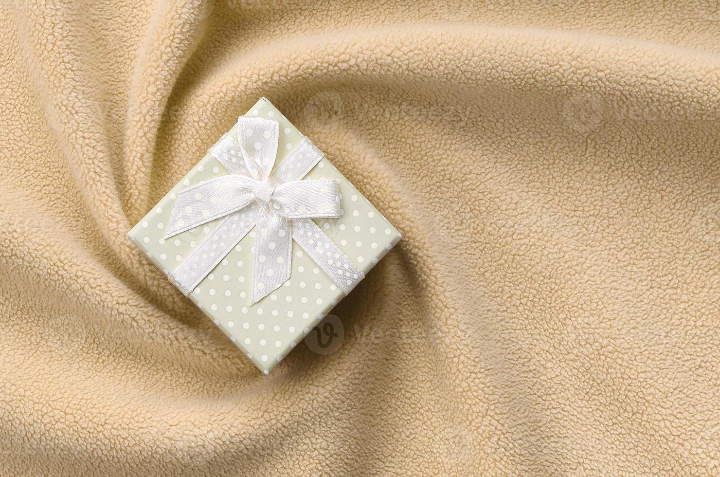 A small gift box in orange with a small bow lies on a blanket of soft and furry light orange fleece fabric with a lot of relief folds. Packing for a gift to your lovely girlfriend photo