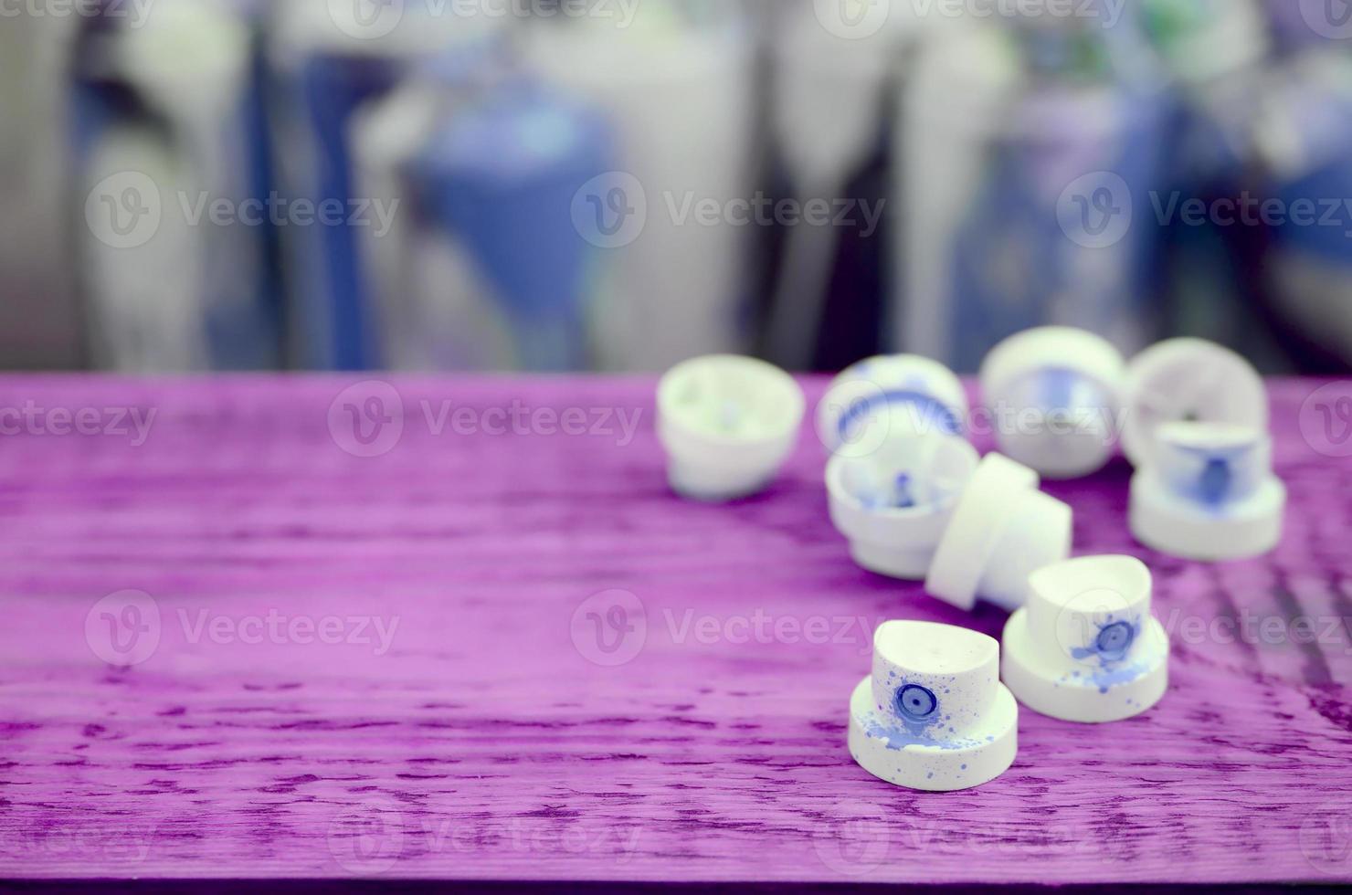 The soiled blue nozzles from the paint sprayer lies on a purple plank on a background of a many dirty spray cans photo