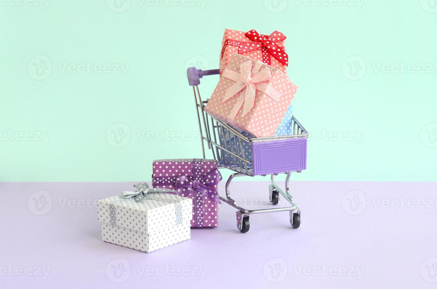 pequeñas cajas de regalo de diferentes colores con cintas se encuentran en el carro de la compra sobre un fondo violeta y azul pastel foto