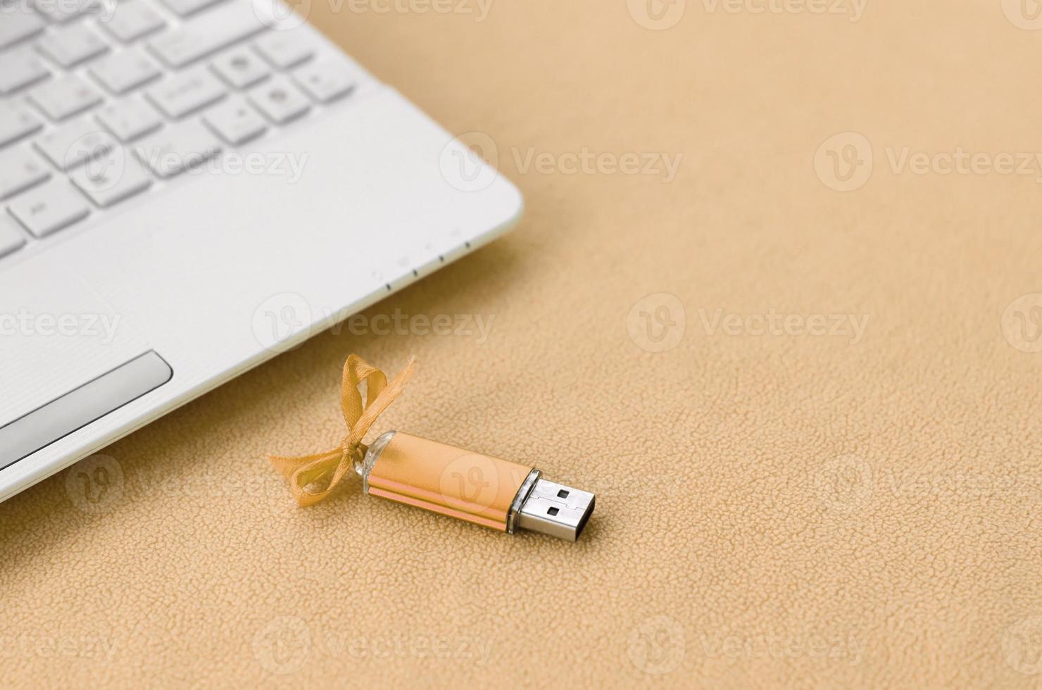 Orange usb flash memory card with a bow lies on a blanket of soft and furry light orange fleece fabric beside to a white laptop. Classic female gift design for a memory card photo