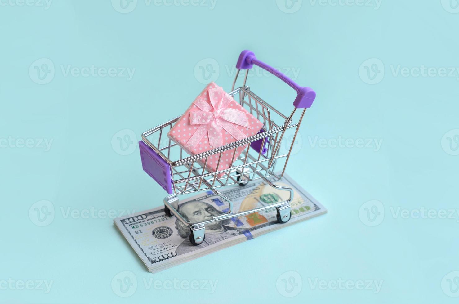 Gift box in a small shopping cart lies on a dollar bills on light blue background photo