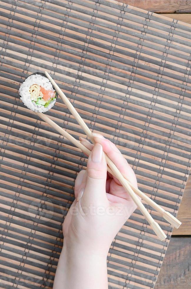 A hand with chopsticks holds a sushi roll on a bamboo straw serwing mat background. Traditional Asian food photo