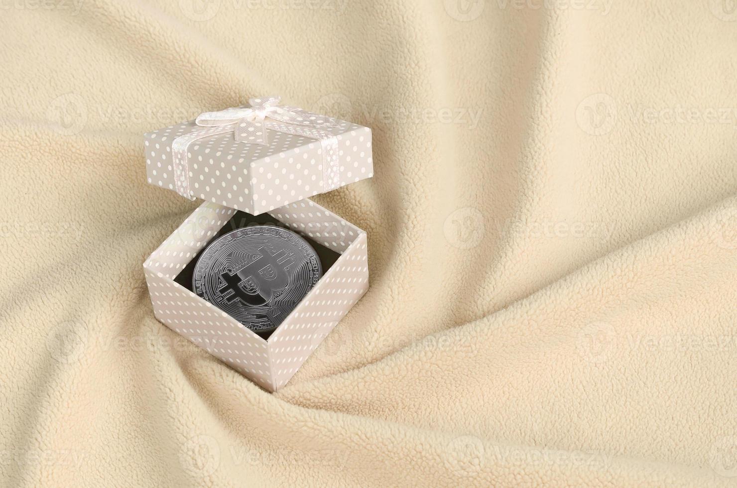 The silver bitcoin lies in a small orange gift box with a small bow on a blanket made of soft and fluffy light orange fleece fabric with a large number of relief folds photo