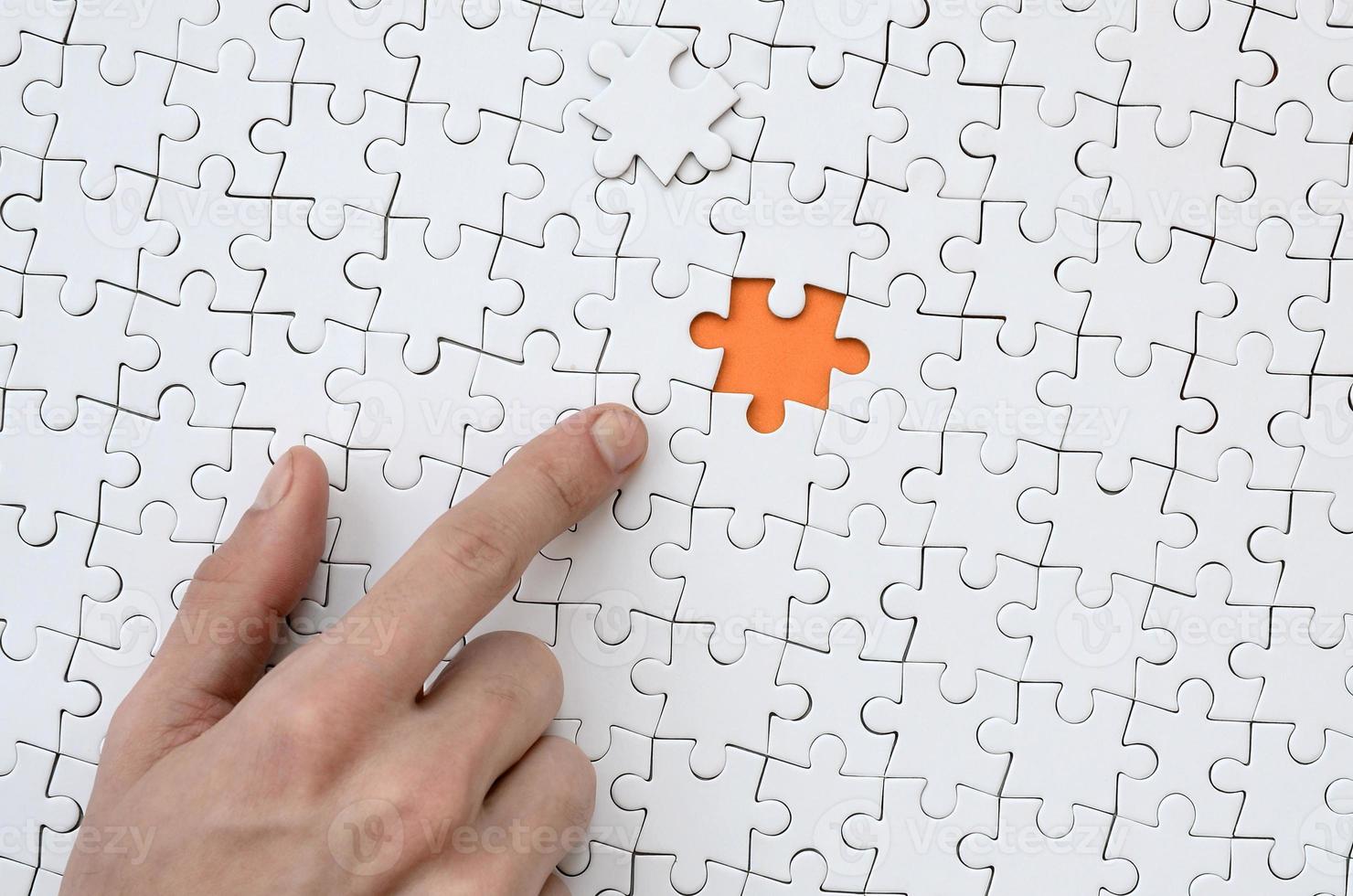 The texture of a white jigsaw puzzle in the assembled state with one missing element, forming an orange space, pointed to by the finger of the male hand photo