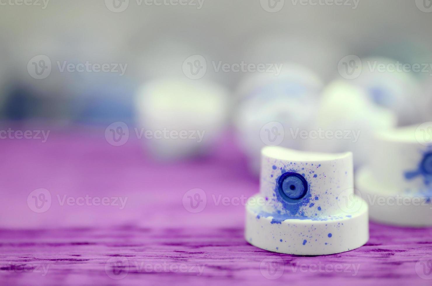 The soiled blue nozzles from the paint sprayer lies on a purple plank on a background of a many dirty spray cans photo
