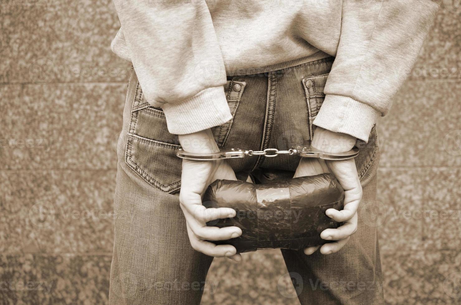 Arrested drug dealer in police handcuffs with big heroin drug package on dark wall background photo