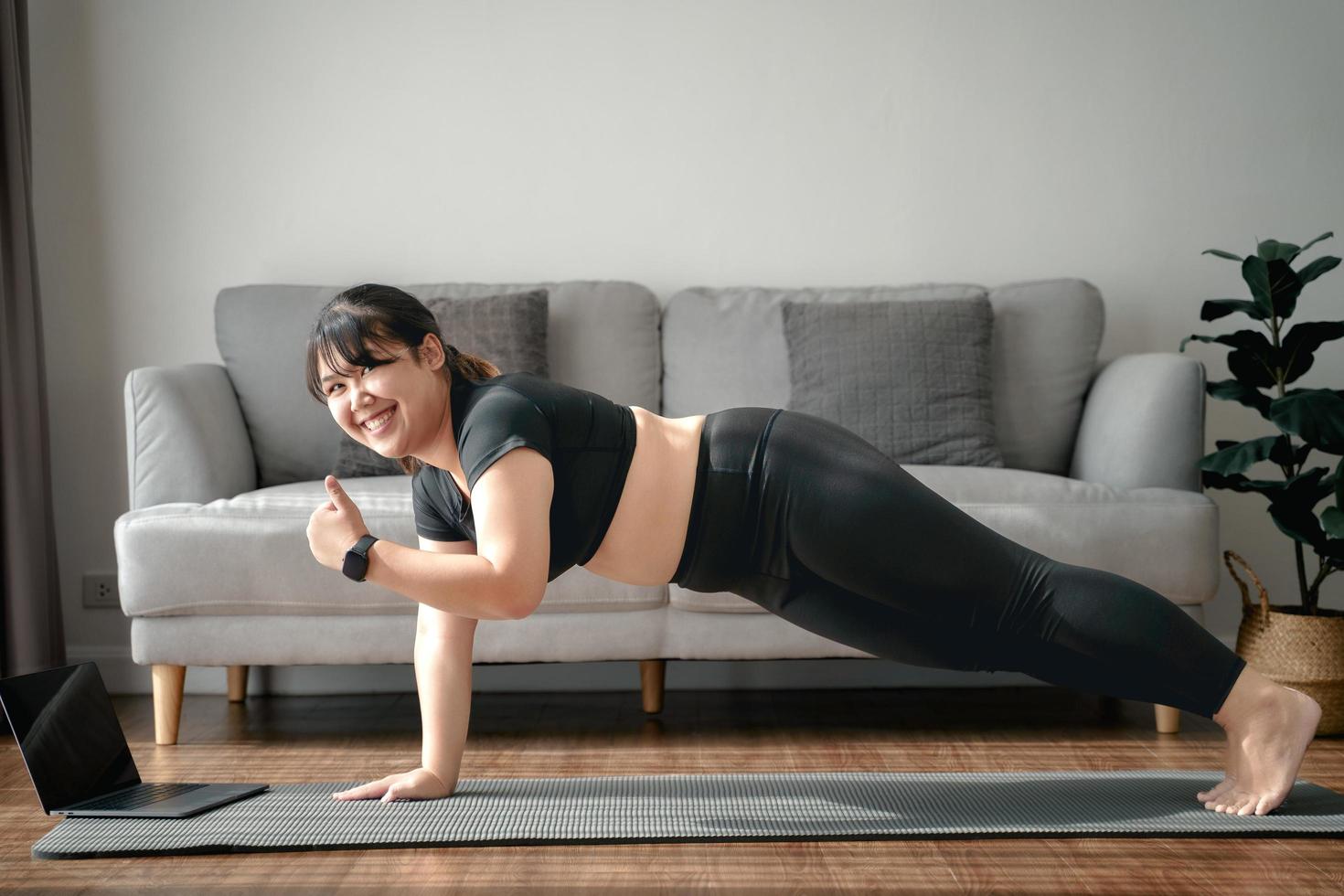 mujer gordita asiática en la sala de estar practica una lección de yoga en línea con la computadora. mujer con clase de entrenamiento de meditación en la computadora portátil. foto