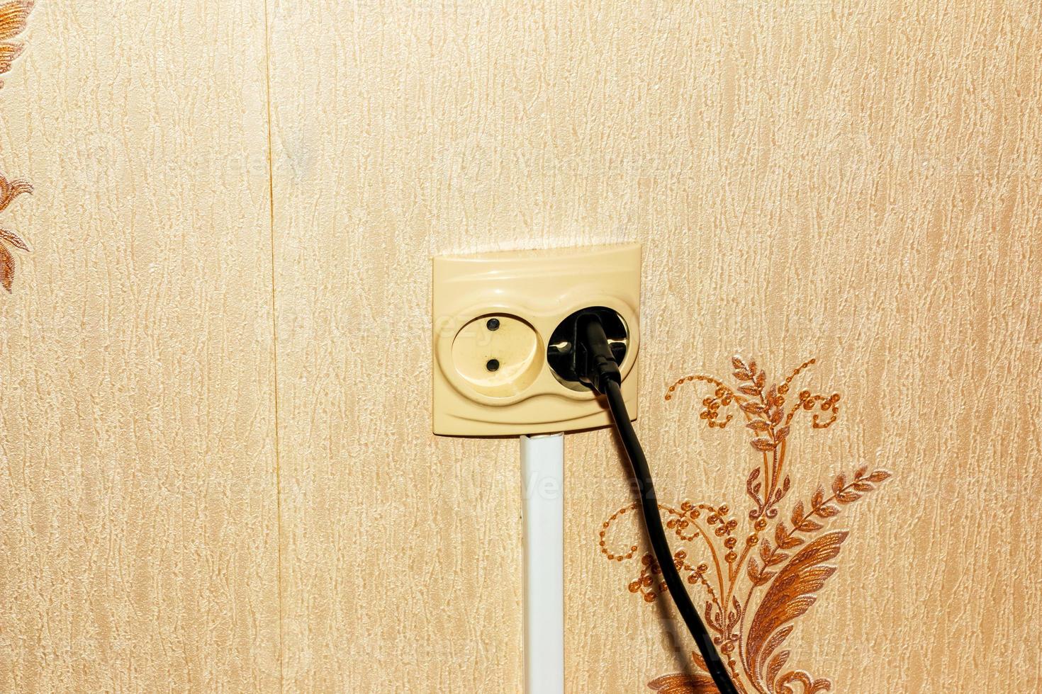 Black electrical cord connected to a plastic white socket on a plain wall. photo