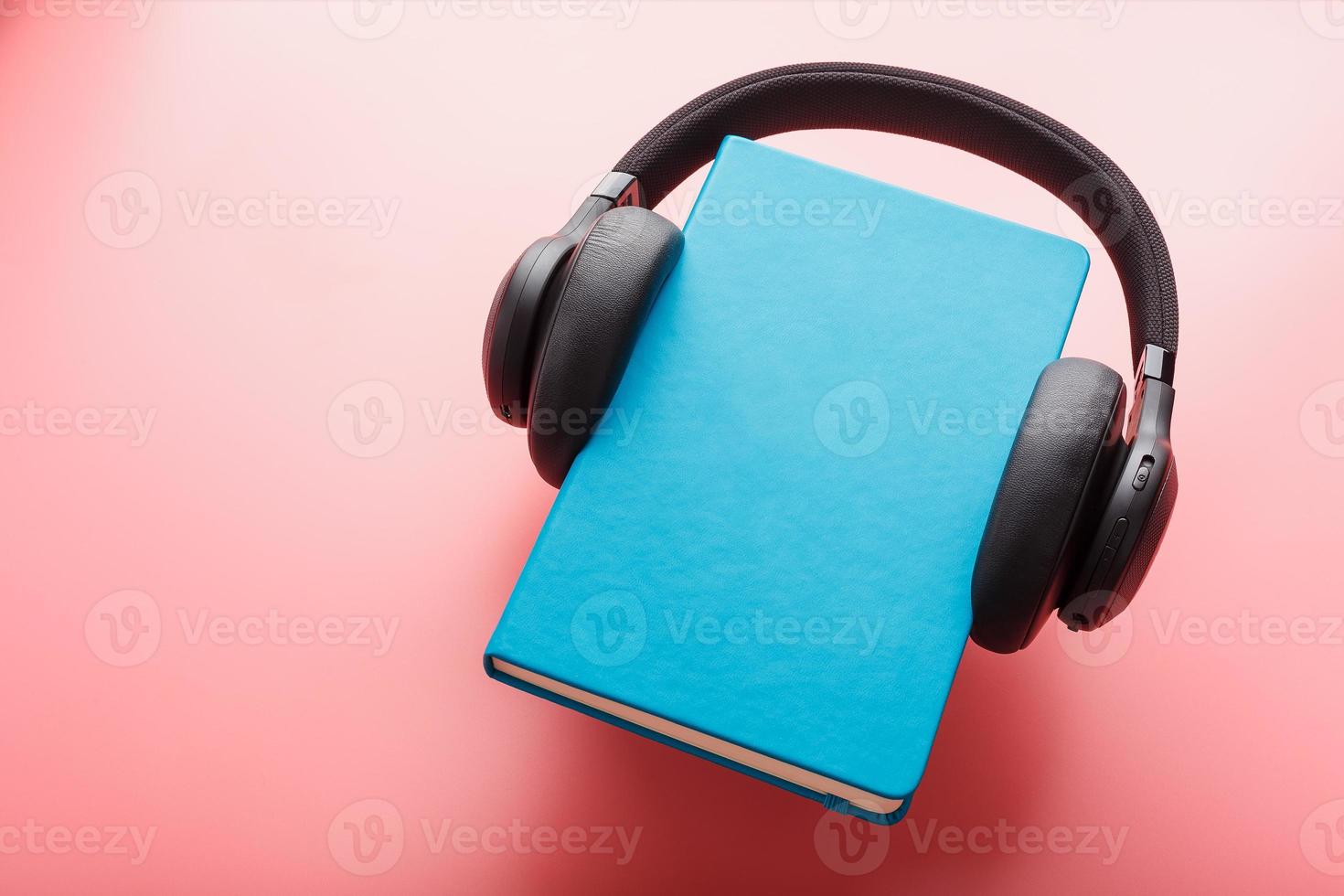 Headphones are worn on a book in a blue hardcover on a pink background, top view. photo