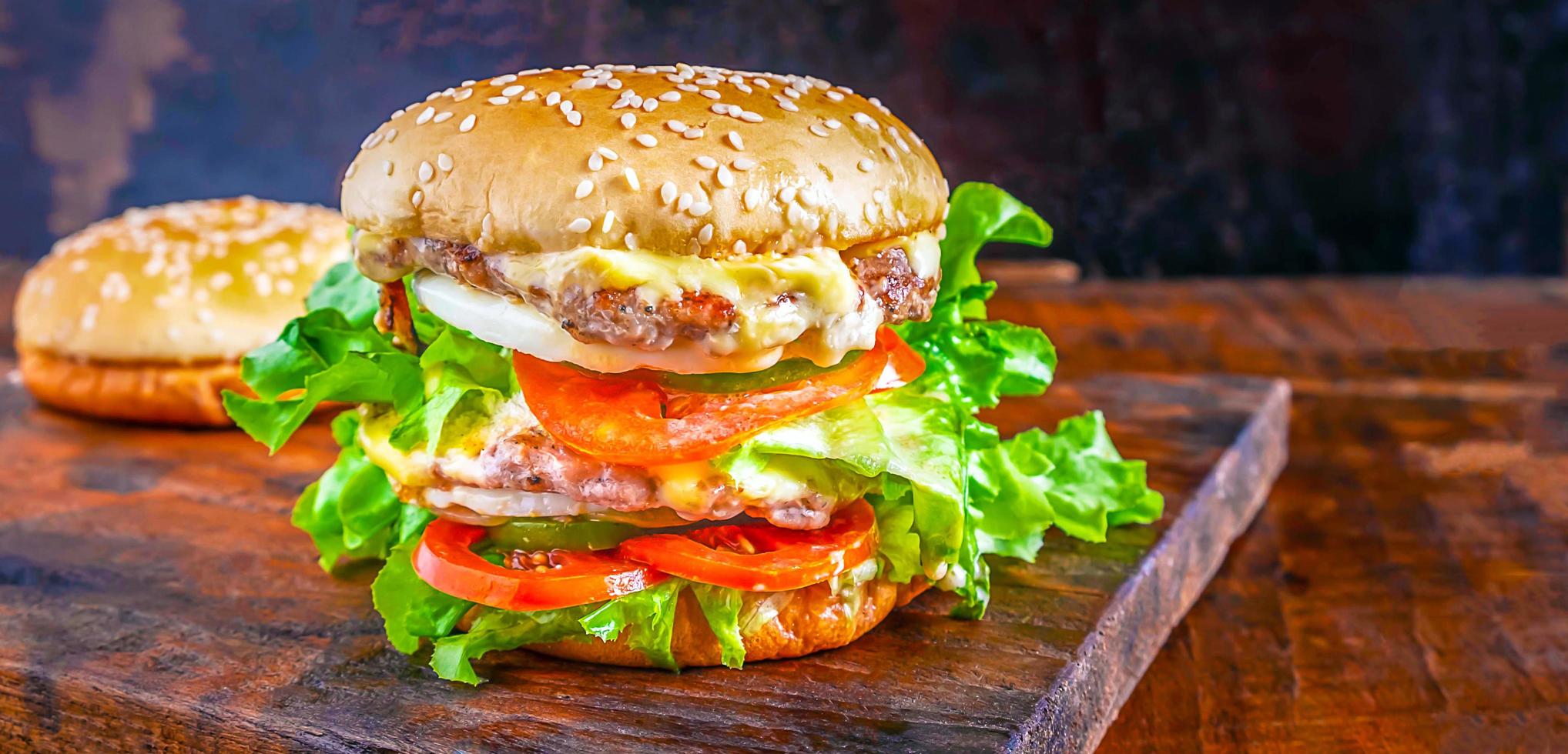 primer plano de deliciosa hamburguesa casera fresca con lechuga, queso, cebolla y tomate en una tabla rústica de madera sobre una mesa de madera foto