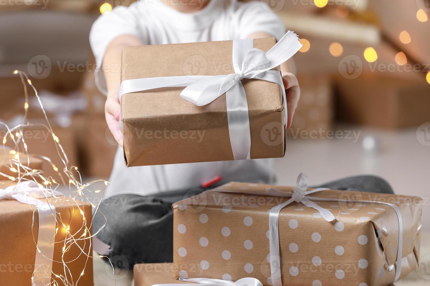 simple gift in kraft paper packaging tied with a white ribbon in the hands of a child boy in a t shirt. baby holding gift box on christmas background. concept of new year, mothers day, Valentines day photo