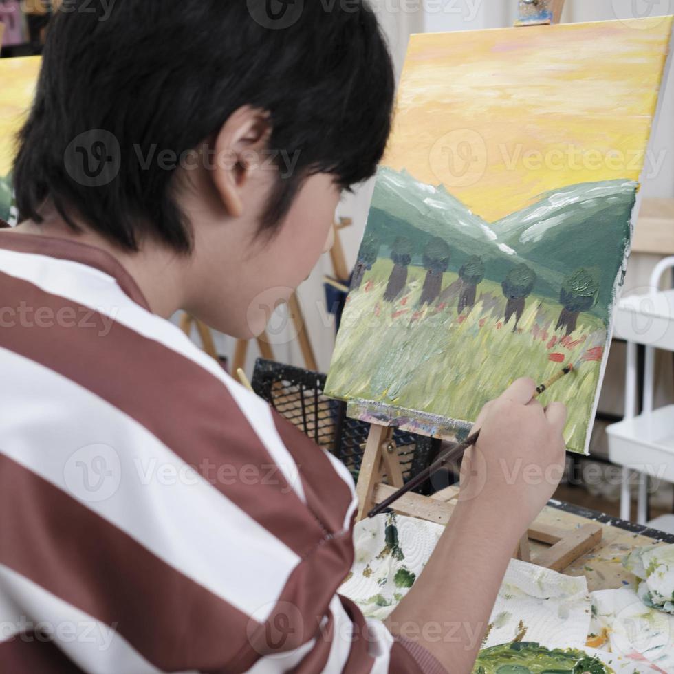 A young Asian boy concentrates on acrylic color picture painting on canvas with student kids in an art classroom, creative learning with talents and skills in the elementary school studio education. photo