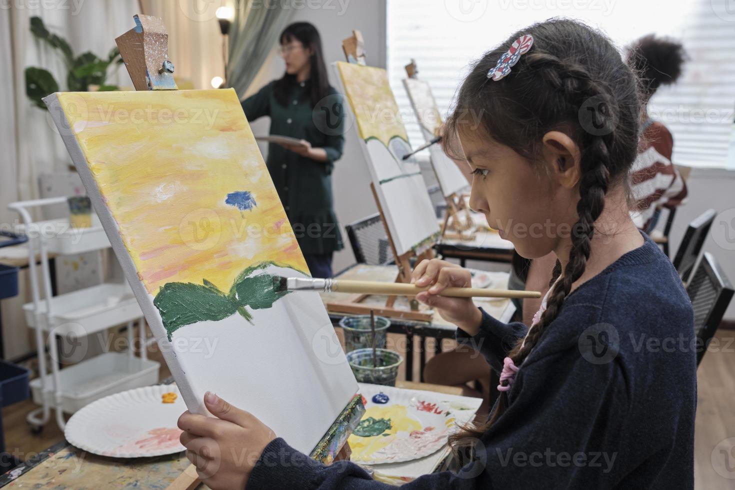 A little girl concentrates on acrylic color picture painting on canvas with student children in an art classroom, creative learning with talents and skills in the elementary school studio education. photo