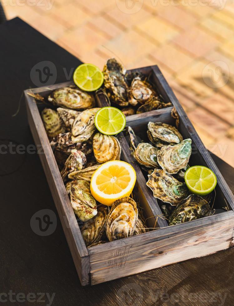 una variedad de ostras frescas con lima y limón en una caja de madera. mariscos frescos terraza de café al aire libre. fondo borroso con vista al club náutico. foto