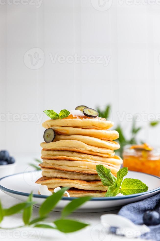 Stack of fluffy pancakes with orange jam, blueberries, coconut chips and mint, light background. Traditional breakfast. High key photography. photo